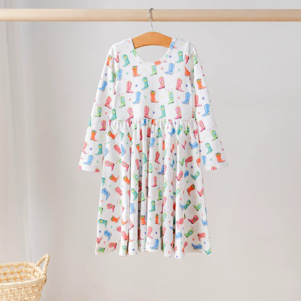 White long-sleeve dress with colorful cowboy boots and stars, a gathered skirt, and a round neckline, hanging on a wooden hanger.